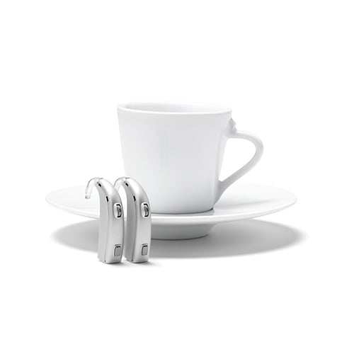 two behind the ear (BTE) hearing aids standing on end in front of a coffee cup on a saucer